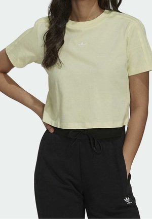 Vrouw die een lichtgeel cropped Adidas T-shirt en zwarte Adidas trainingsbroek draagt met handen in de zij, bij de nek en taille bijgesneden.