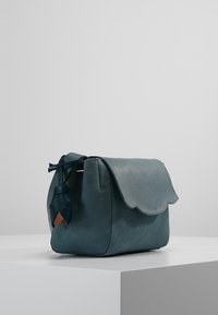 Tealfärgad läderväska med en scallopformad lock, texturerad yta och en accenttassel i en mörkare nyans. Har en strukturerad form och minimalistisk design.