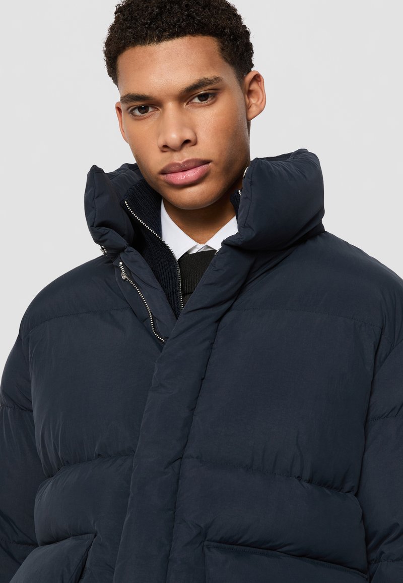 Veste matelassée marine avec un col montant, texture matelassée, fermeture éclair à l'avant et poches latérales. Portée sur une chemise blanche et une cravate.