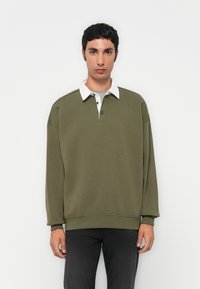 Olivgrüner Pullover mit weißem Kragen und grauem Innenfutter. Hat eine lockere Passform und gerippte Bündchen. Zwei Druckknöpfe im Halsbereich.