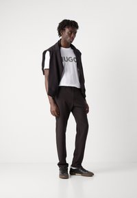 Black sweater draped over shoulders, white T-shirt with "HUGO" graphic, brown trousers, and black sneakers with gray accents.