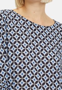 Blouse à motifs bleu et blanc présentant des formes circulaires et en losange, sur un fond marine, avec une texture lisse et un décolleté en scoop.