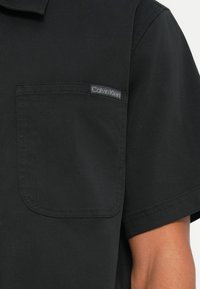 Črna kratka majica s kratkimi rukavi z žepom na prsih, ki vsebuje oznako Calvin Klein, izdelana iz gladkega materiala s čistim in preprostim dizajnom.