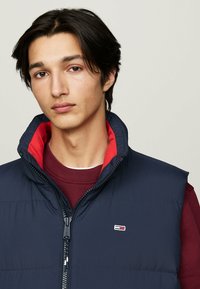 Navy sleeveless puffer vest with red lining, zippered front, and small logo on the chest. Worn over a burgundy sweater. Smooth texture.