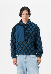 Fleece jacket featuring a blue base with a textured black geometric pattern, zip closure, and side pockets. Soft, cozy feel.