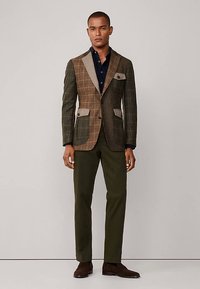 Karierter Stoffblazer in Braun- und Grüntönen, kontrastreiche beigen Revers, Vordertaschen mit Klappen, kombiniert mit dunklem Hemd und olivgrüner Hose.