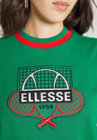 Femme portant un sweat-shirt vert avec un col rouge et des raquettes de tennis brodées, un filet et le logo "ELLESSE 1959" à l'avant.