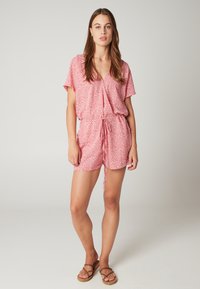 Rode bloemen romper met korte mouwen, V-hals, drawstring taille en relaxte pasvorm. Draagt met bruine sandalen en minimale accessoires.