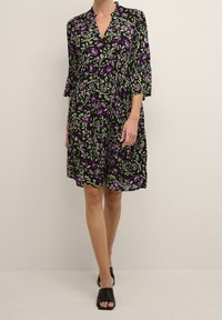 Robe florale noire avec des motifs violets et verts, décolleté en V, manches trois-quarts et coupe ample. Portée avec des chaussures noires ouvertes.
