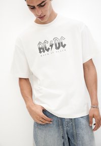 Bílý bavlněný tričko s grafickým potiskem "AC/DC" a "BACK IN BLACK" v černých písmenech. Krátké rukávy a pohodlný střih kalhot.