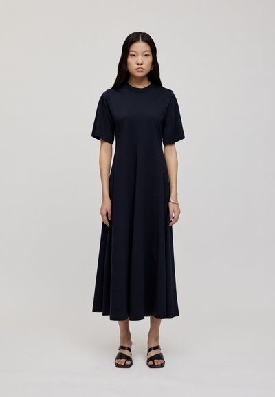 Granatowa sukienka z krótkim rękawem o okrągłym dekolcie, płynącym kształcie w literę A i miękkiej, gładkiej teksturze. Zestawiona z czarnymi sandałami na paski.