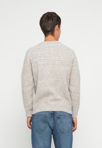 Homme de dos portant un pull texturé gris clair et un jean bleu avec poches arrière, contre un fond blanc uni.
