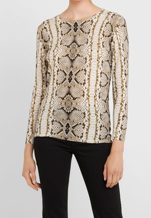 Femme portant un haut ajusté à manches longues avec un motif peau de serpent beige et marron, associé à un pantalon noir, sur un fond gris clair.