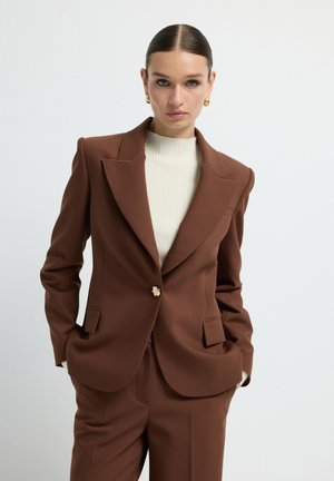 Blazer marron ajusté avec des revers nets, fermeture à un bouton et poches latérales, associé à un haut crème c�ôtelé. Texture lisse.