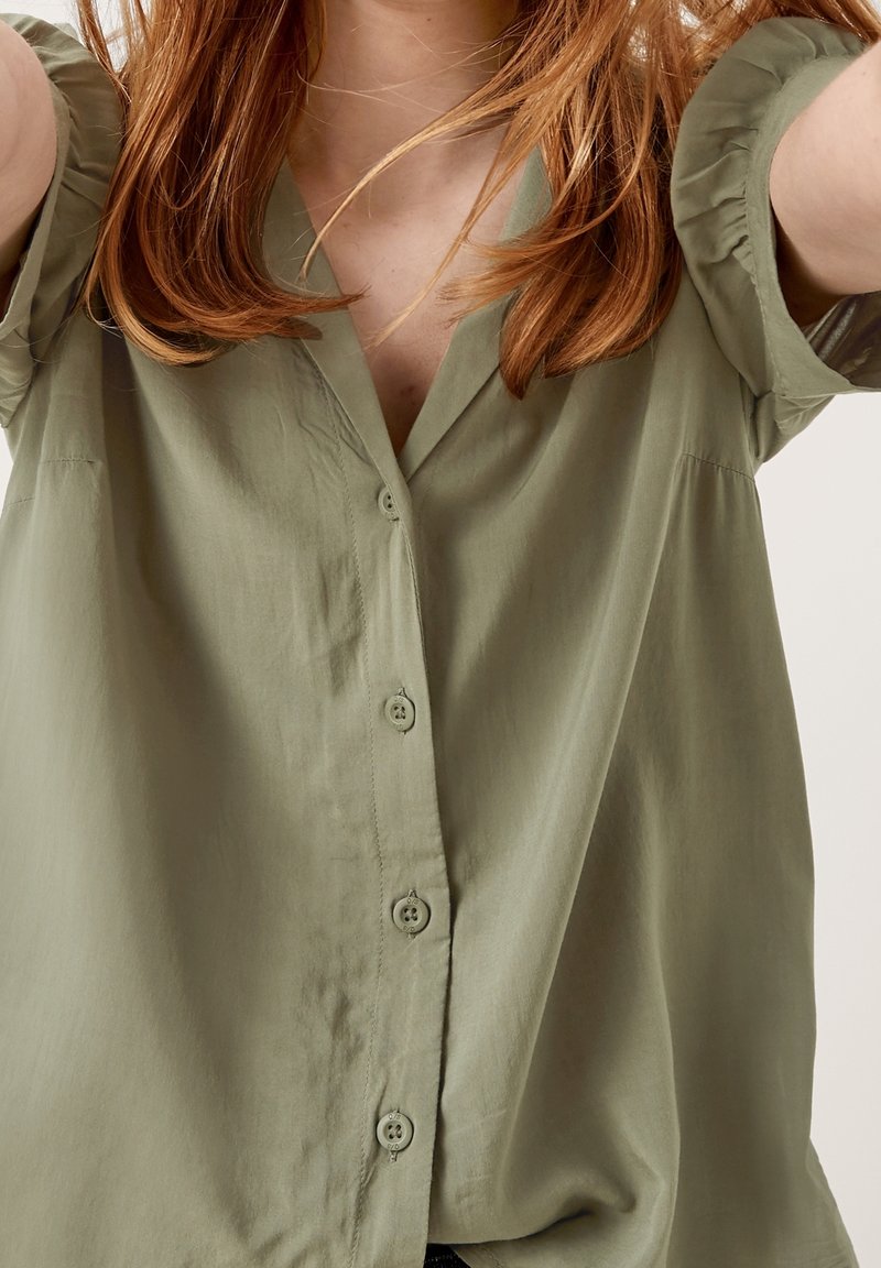 Chemise à manches courtes, de couleur vert olive, à boutons et fabriquée en tissu doux. Elle présente une coupe décontractée, un décolleté en V et une texture subtile.