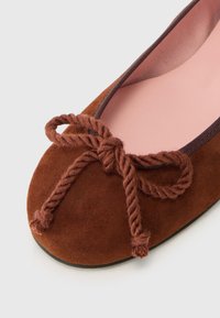 Ballerines en daim marron avec une doublure rose et un détail de nœud en corde tressée à l'orteil, présentant une forme arrondie et une semelle plate.