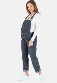 Donkergrijze denim tuinbroek. Kenmerkt zich door verstelbare schouderbanden, een voorzak en opgerolde mouwen. Draagt over een aansluitend wit lange mouwen top.