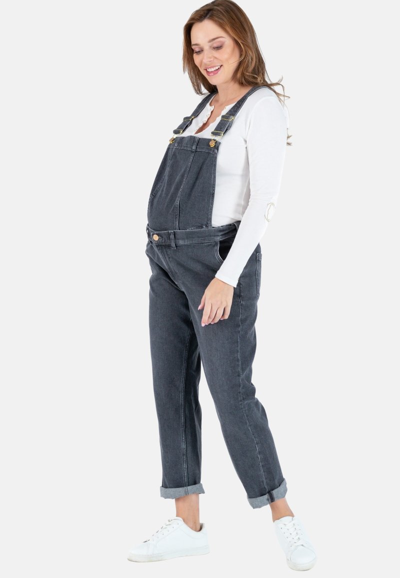 Donkergrijze denim tuinbroek. Kenmerkt zich door verstelbare schouderbanden, een voorzak en opgerolde mouwen. Draagt over een aansluitend wit lange mouwen top.