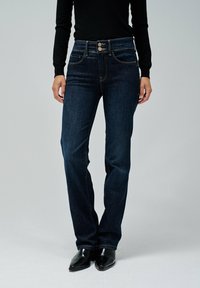 Jeans de mezclilla azul oscuro con cintura alta, dos botones, cinco bolsillos y detalles de costura sutiles. La modelo lleva botas negras hasta el tobillo.
