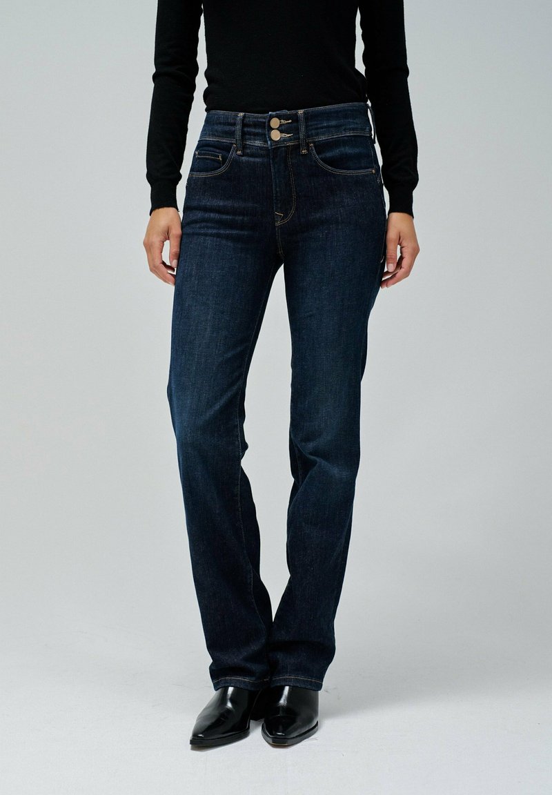 Jeans de mezclilla azul oscuro con cintura alta, dos botones, cinco bolsillos y detalles de costura sutiles. La modelo lleva botas negras hasta el tobillo.