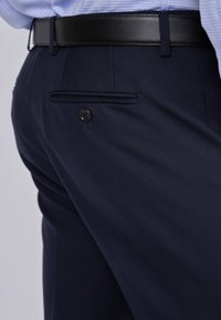 Pantalons navy en tissu texturé, dotés d'une poche arrière à boutons et d'une ceinture en cuir noir. La coupe ajustée met en valeur un style contemporain.