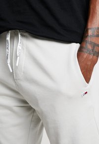 Primer plano de una persona que lleva pantalones Tommy Jeans en color blanco roto, con el logotipo visible en el bolsillo, cordones con texto de la marca y un tatuaje en el brazo descansando en el bolsillo.