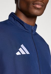 Μπλε ναυτικού χρώματος ζακέτα με φερμουάρ, που διαθέτει ψηλό γιακά και λευκό έμβλημα με τρεις ρίγες στο αριστερό μέρος του στήθους, κατασκευασμένη από λείο, ελαφρύ ύφασμα.