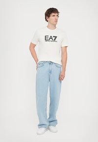 White cotton t-shirt with black "EA7 Emporio Armani" logo, paired with light blue denim jeans. Sneakers are white with textured soles.