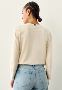 Beige pitkähihainen paita pyöreällä pääntiellä, tekstuurillisesta kankaasta ja takana tummansininen merkki, yhdistetty vaaleansinisiin korkeavyötäröisiin farkkuihin.