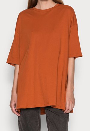 Persona con una camiseta holgada de manga corta color naranja quemado y pantalones gris oscuro, de pie frente a un fondo blanco liso.