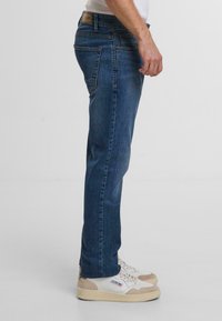 Donkerblauwe denim jeans met een rechte pasvorm, voorzien van vijf zakken en subtiele stikdetails, gecombineerd met witte en beige sneakers.