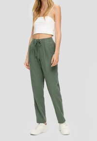 Leichte grüne Leinenhosen mit einem Kordelzugbund, schmal zulaufenden Beinen und seitlichen Taschen, kombiniert mit einem weißen, kurz geschnittenen Tanktop und weißen Sneakers.