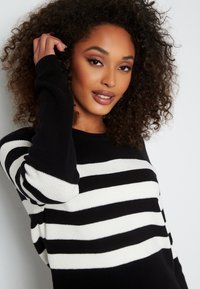 Maglione a righe bianche e nere con scollatura rotonda, morbido tessuto a maglia e maniche lunghe. Capelli ricci coprono parzialmente una mano.