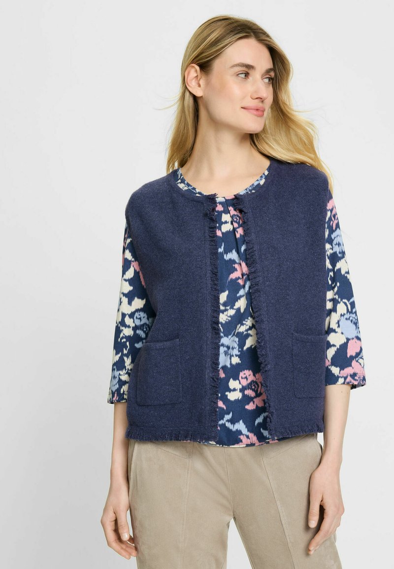 Vrouw in beige broek, marineblauwe bloes met bloemenpatroon en mouwloos marineblauw vest met zakken aan de voorkant, kijkend naar de zijkant tegen een effen achtergrond.