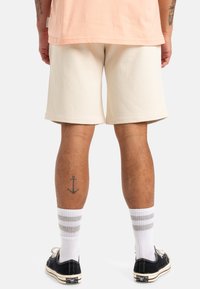 Pantalones cortos de algodón beige con un ajuste holgado, que llegan por encima de la rodilla, combinados con calcetas blancas con rayas grises y zapatillas negras.