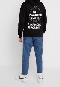 Černá mikina s kapucí a bílým textem na zádech: „GET SOMETHING CLEVER“ a „A DIAMOND IS FOREVER.“ Džínové kalhoty a bílé tenisky.