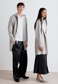 Deux modèles se tiennent côte à côte. Le homme porte un imperméable gris clair à boutons, un t-shirt blanc et un pantalon noir. La femme porte un manteau similaire sur un haut blanc et une jupe noire, avec des talons argentés.