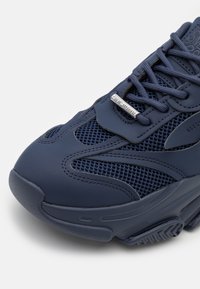 Zapatilla navy que presenta una combinación de texturas lisas y de malla, con punta redondeada, cordones y un acento metálico de marca en la parte superior.