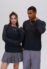 Maglioncini a maglia blu navy con un design a rete aperta. La modella femminile indossa una gonna grigia plissettata, e entrambe le modelle portano collane di catena argentata.