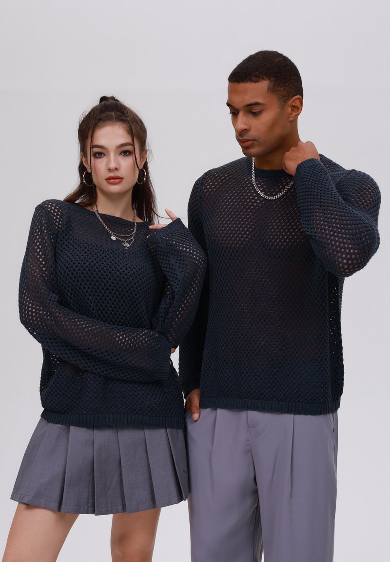 Maglioncini a maglia blu navy con un design a rete aperta. La modella femminile indossa una gonna grigia plissettata, e entrambe le modelle portano collane di catena argentata.