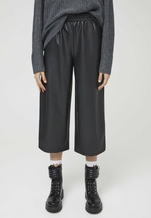 Culottes en simili cuir noir avec une taille élastique, associées à des bottes de combat noires avec un poignet côtelé, sur un fond neutre.