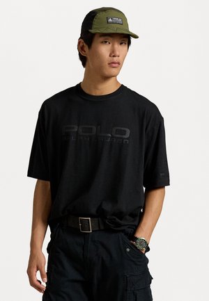 Camiseta negra de gran tamaño con un logo texturizado de "POLO RALPH LAUREN", combinada con una gorra verde y negra con un parche de logo, y pantalones cargo.
