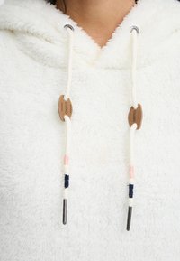 Balta pūkinė džemperis su tekstūrizuota paviršiumi. Šnūrų su natūralaus medžio užtrauktuku ir metaliniais galais akcentuoja šviesiai rožinės ir tamsiai mėlynos spalvos siūlai.