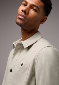 Camisa de botones de color gris claro, confeccionada con una tela suave, que presenta un cuello, un bolsillo con cierre de botón y una textura sutil.