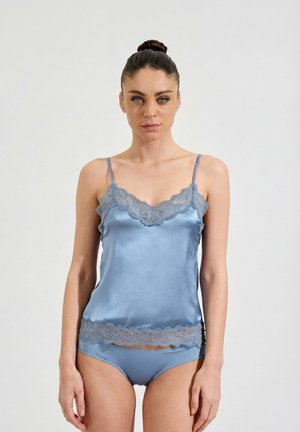 Frau mit Haarknoten trägt hellblaues Satin-Camisole und passende Unterwäsche mit Spitzenbesatz, stehend vor schlichtem hellem Hintergrund.