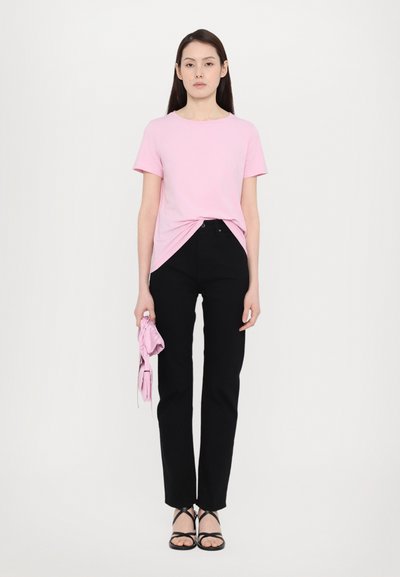 Rosa bomull t-skjorte med korte ermer og knute-detalj, sammen med svarte rette jeans og svarte sandaler med stropp. Holder en rosa gjenstand.