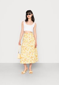 Top cropped branco, saia maxi floral amarela com margaridas, mules amarelos e uma pequena bolsa amarela. Textura suave, ajuste descontraído.