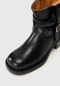 Bottine en cuir noir avec un bout rond, un dessus plis, et un détail de boucle sur le côté. Elle présente un talon bloc solide et une texture lisse.