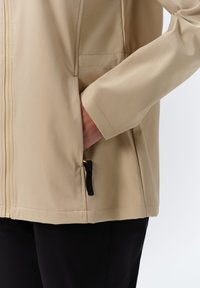Beige softshell jas met een zachte textuur, ritszakken en een getailleerd ontwerp; heeft een grijze rits en zwarte trekaccenten.