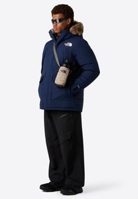 Parka isolata blu navy con cappuccio foderato in pelliccia, tasche laterali e logo. Abbinata a pantaloni neri e porta una borsa beige con una bottiglia d'acqua.
